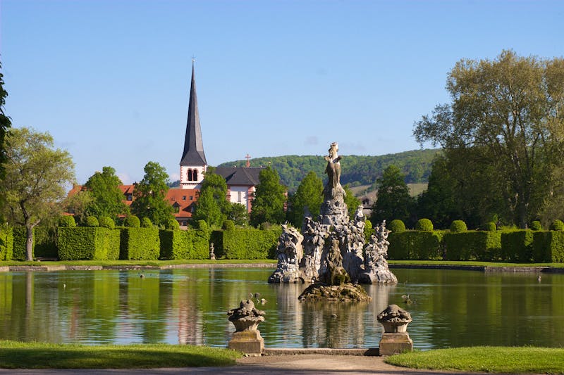 Der Musenberg - Parnassbrunnen im Hofgarten von Veitsöchheim. Die Kirchen t. Vitus im Hintergrund - ©WernerHilpert - stock.adobe.com